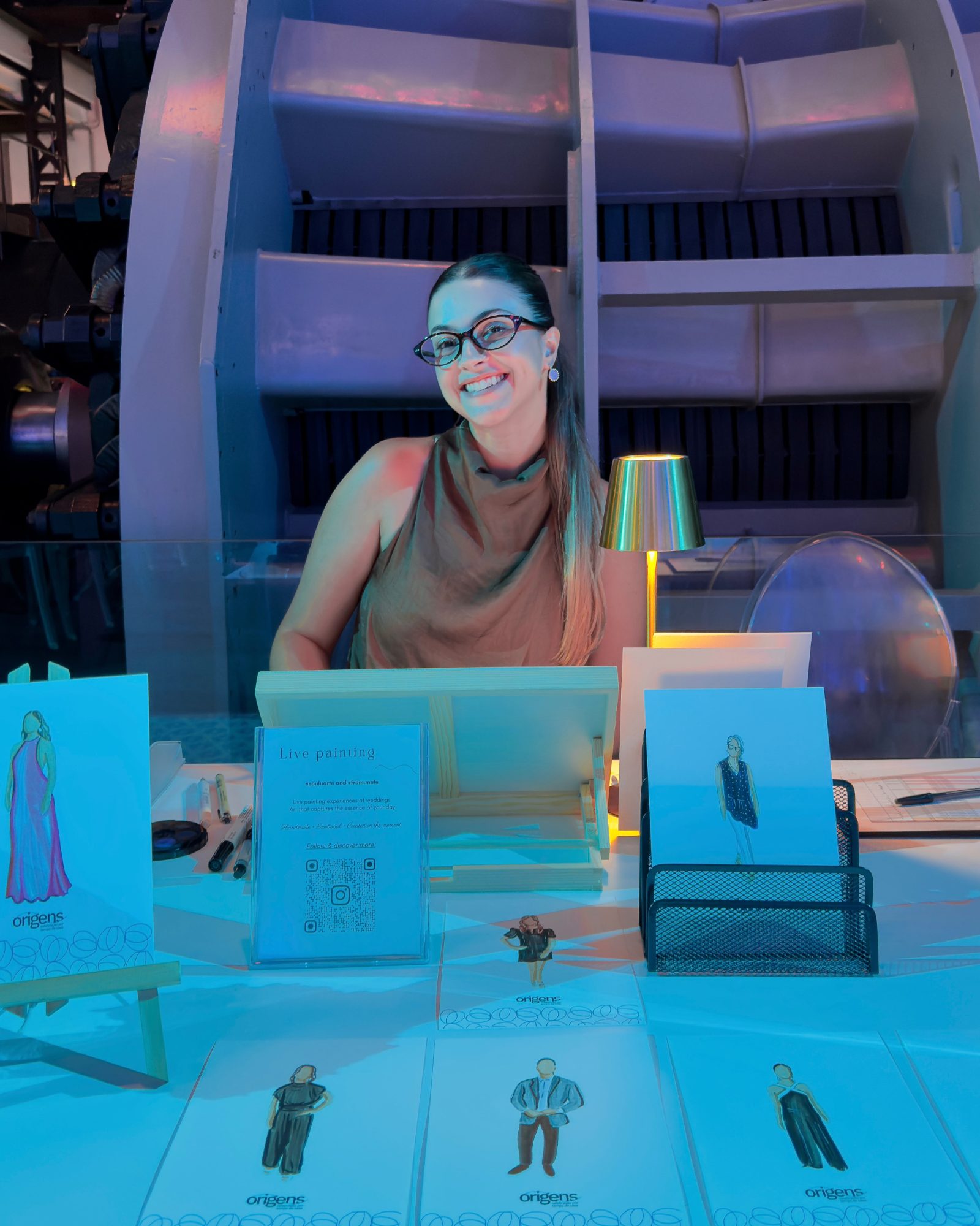 Luíza sorrindo na sua mesa de pintura ao vivo no MAAT, com retratos em aquarela expostos à frente · Souluarte