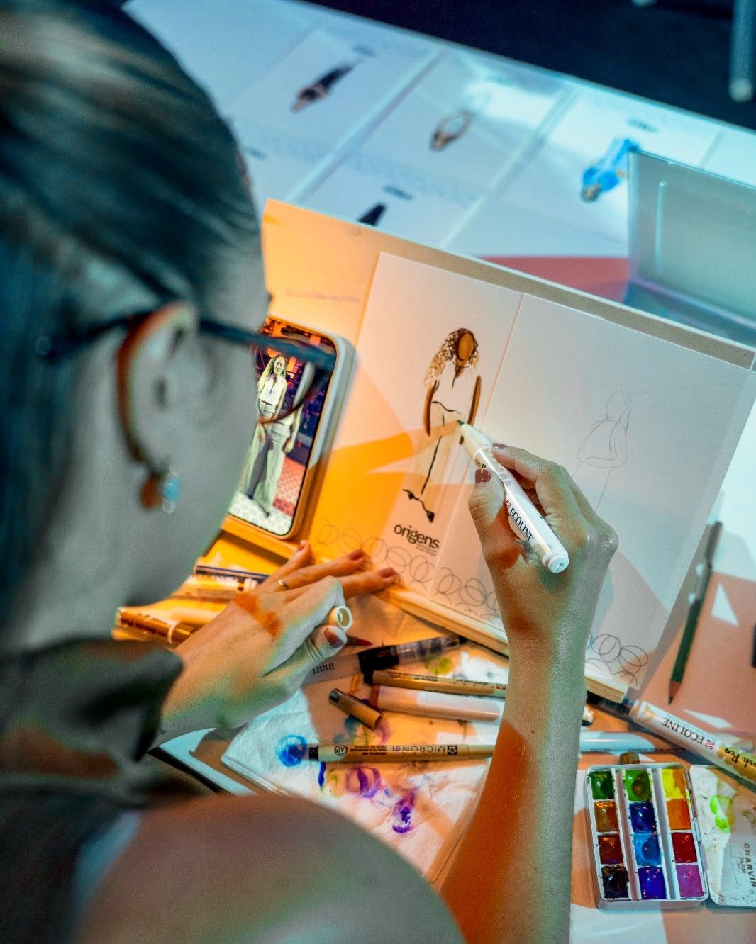 Luíza finaliza um retrato com caneta, rodeada de canetas e aquarelas · Souluarte