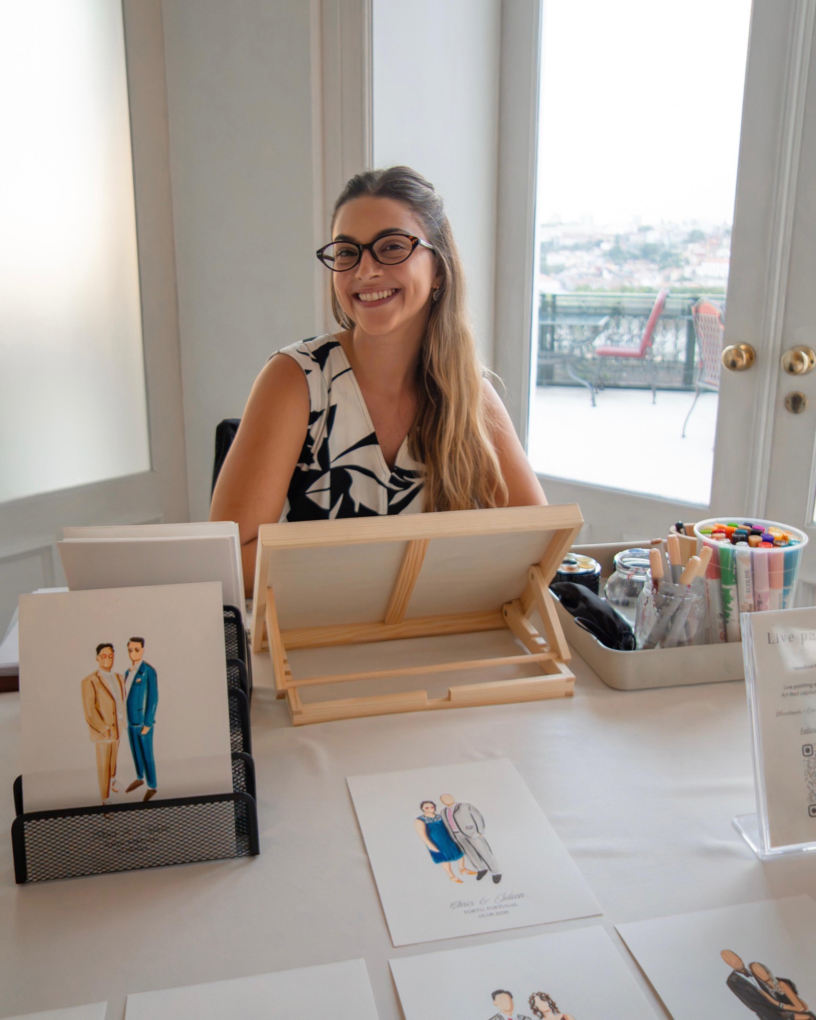 Luíza at her live painting table by the window, portraits and pens ready · Souluarte