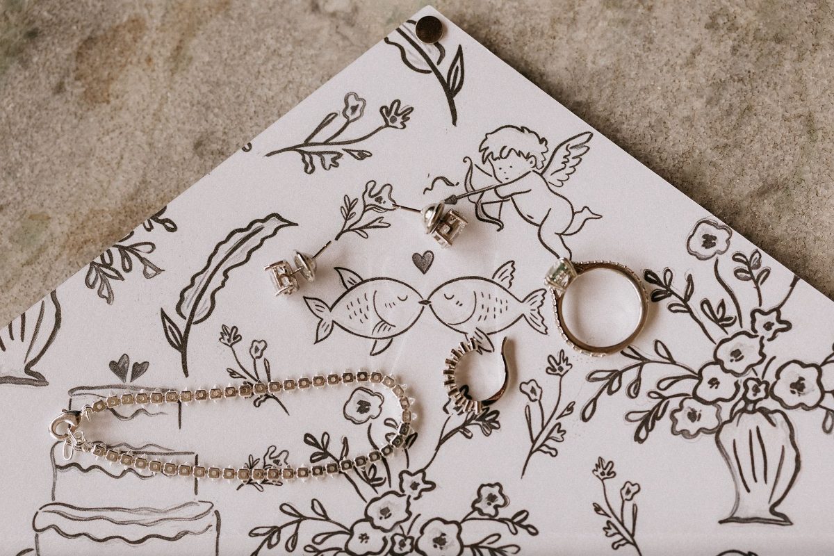 Hand-drawn envelope liner with kissing fish and cupid, with the wedding rings and jewellery laid on top · Souluarte